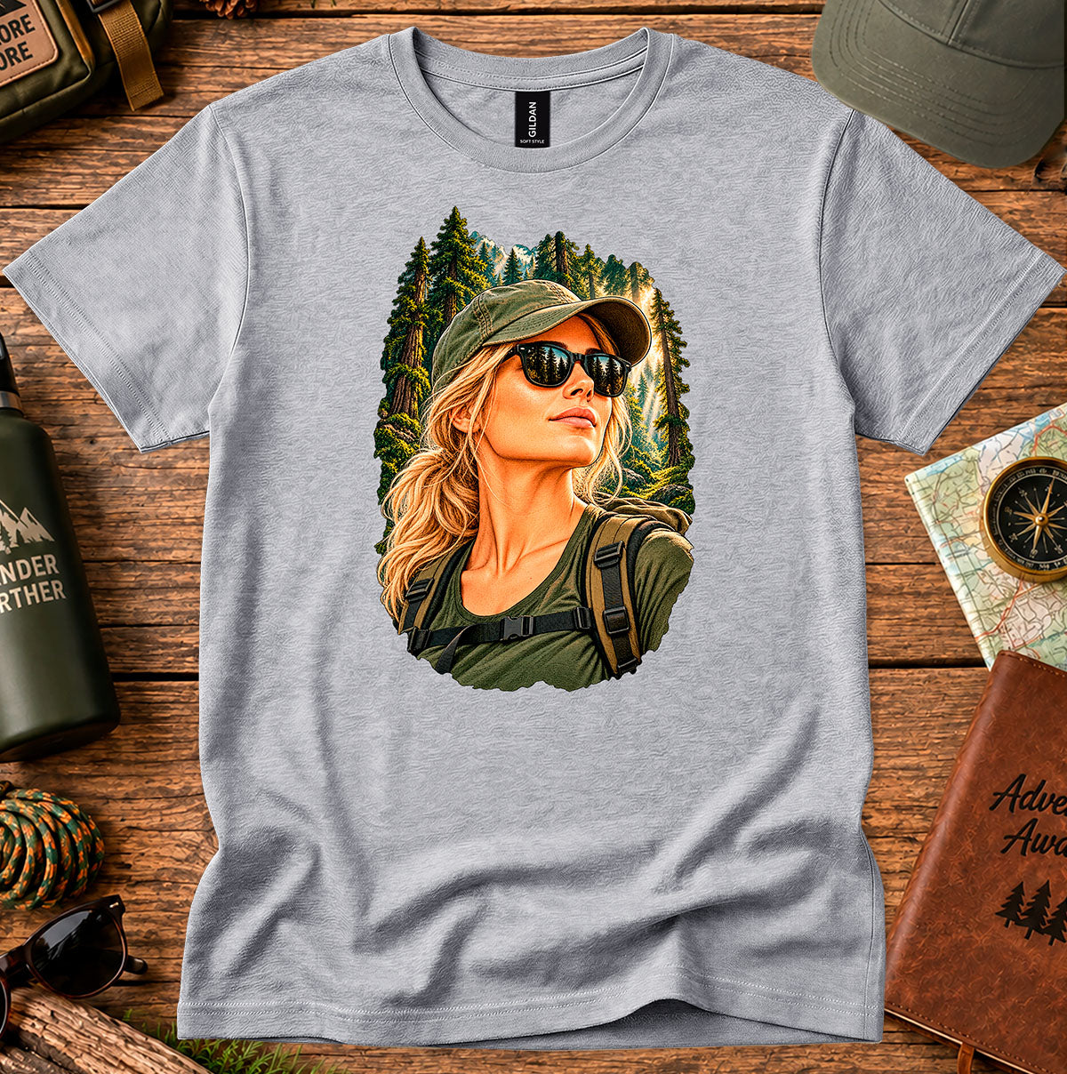 Grey T-shirt featuring a woman wearing a cap and sunglasses in front of evergreen trees