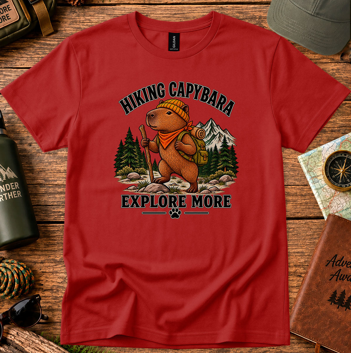 Explore More Hiking Capybara graphic T-shirt featuring a capybara with a backpack and bandana, pine trees and mountains