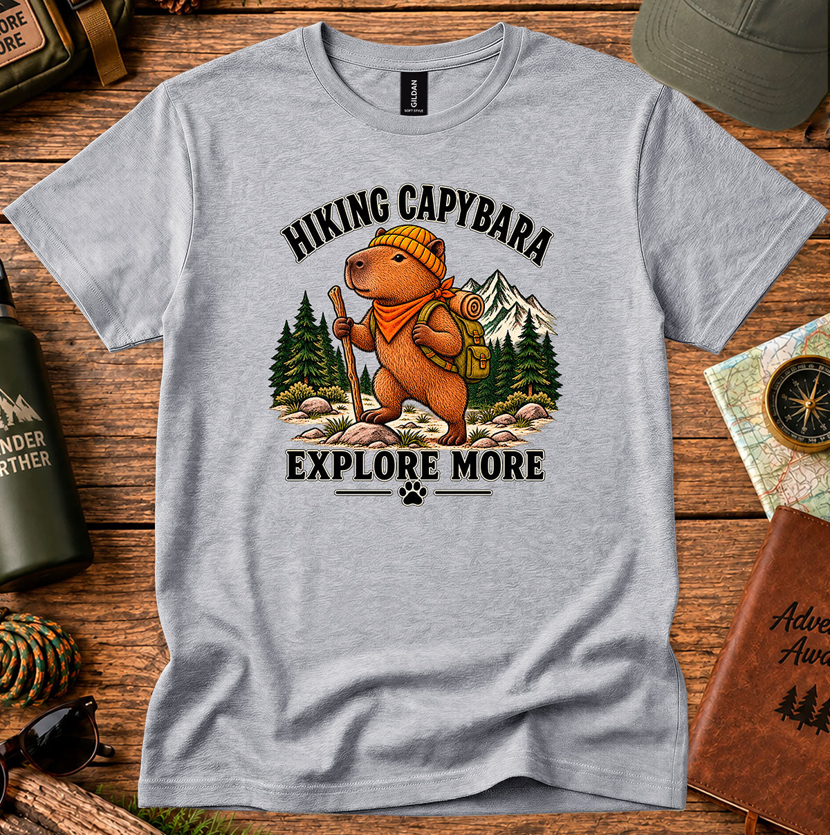 Explore More Hiking Capybara graphic T-shirt featuring a capybara with a backpack and bandana, pine trees and mountains
