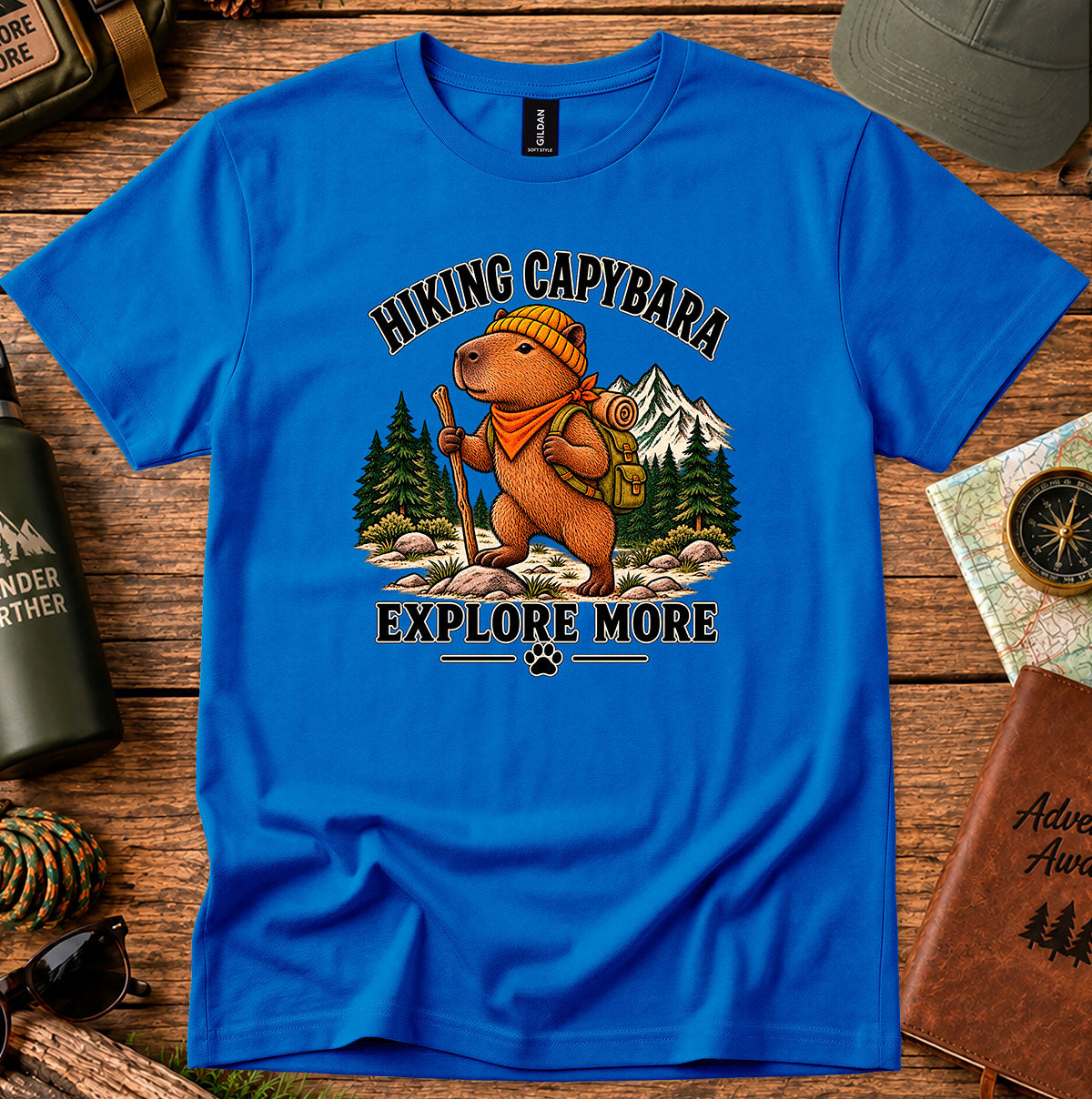 Explore More Hiking Capybara graphic T-shirt featuring a capybara with a backpack and bandana, pine trees and mountains